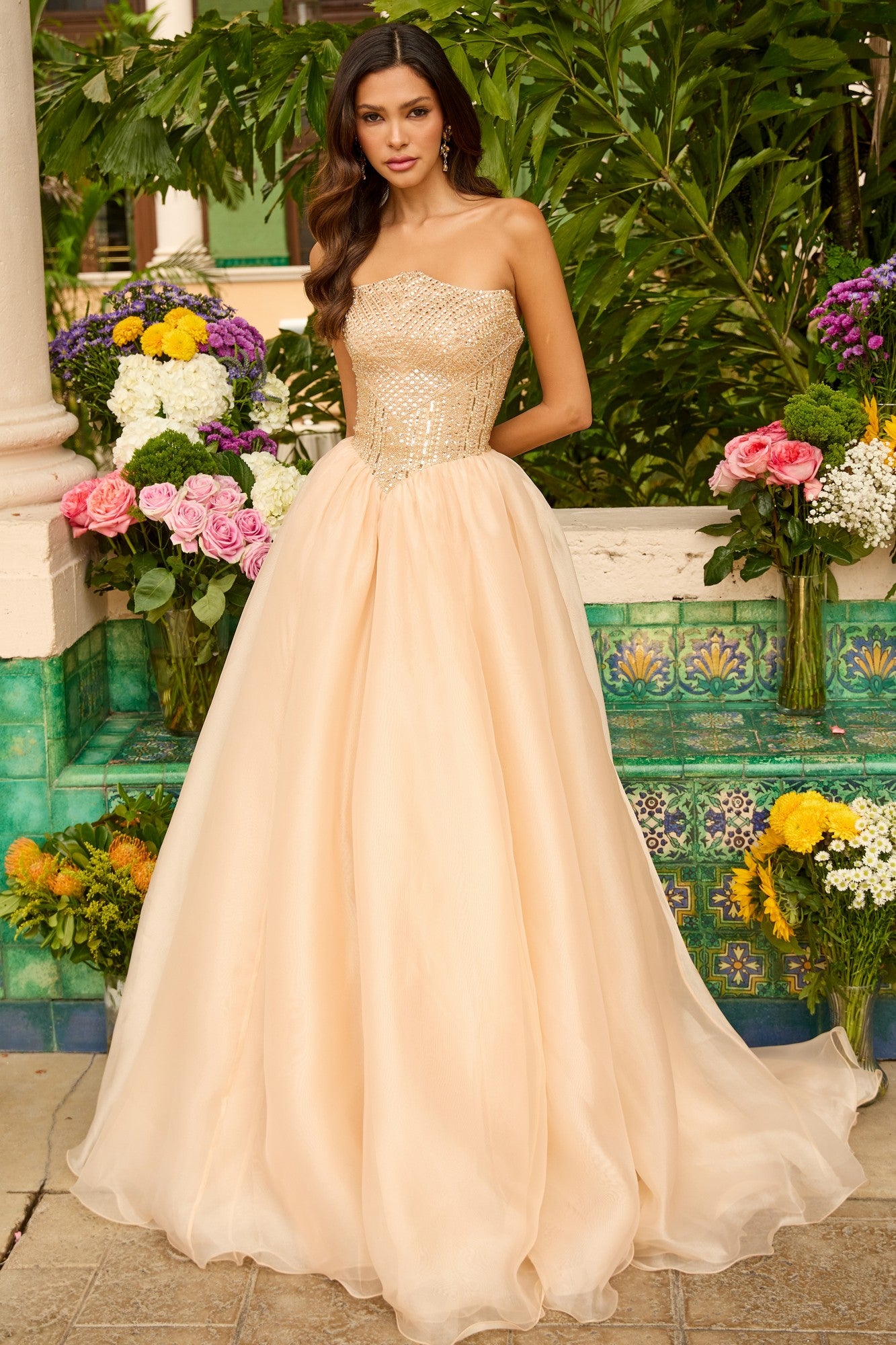 A woman in a gold Ava Presley 47255 long prom dress with a strapless, embellished bodice and full skirt stands outdoors by vibrant flowers and decorative tiles.